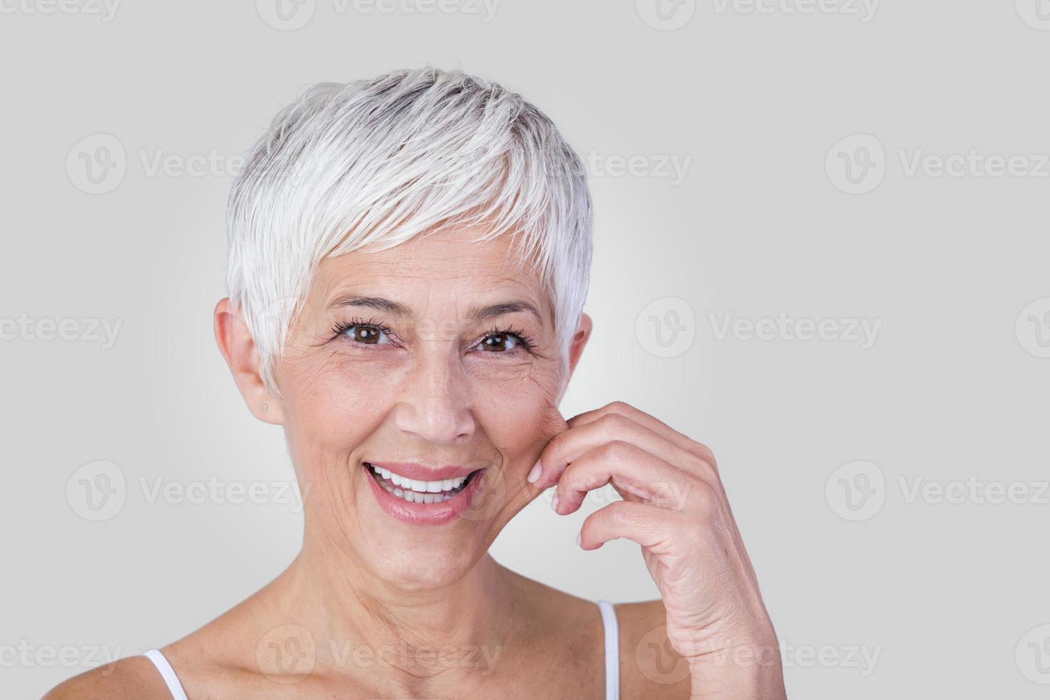 anziano donna traino guance per sentire morbidezza e guardare a telecamera. bellezza ritratto di contento maturo donna sorridente con mani su guancia isolato al di sopra di grigio sfondo. invecchiamento processi e pelle concetto. foto