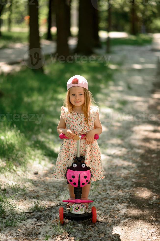 un' poco ragazza 3 anni vecchio nel un' rosa berretto cavalcate un' scooter. estate volta. foto