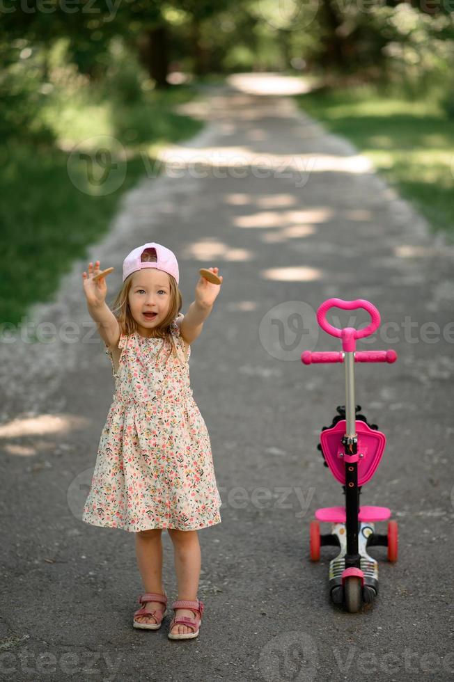 un' poco ragazza 3 anni vecchio nel un' rosa berretto cavalcate un' scooter. estate volta. foto