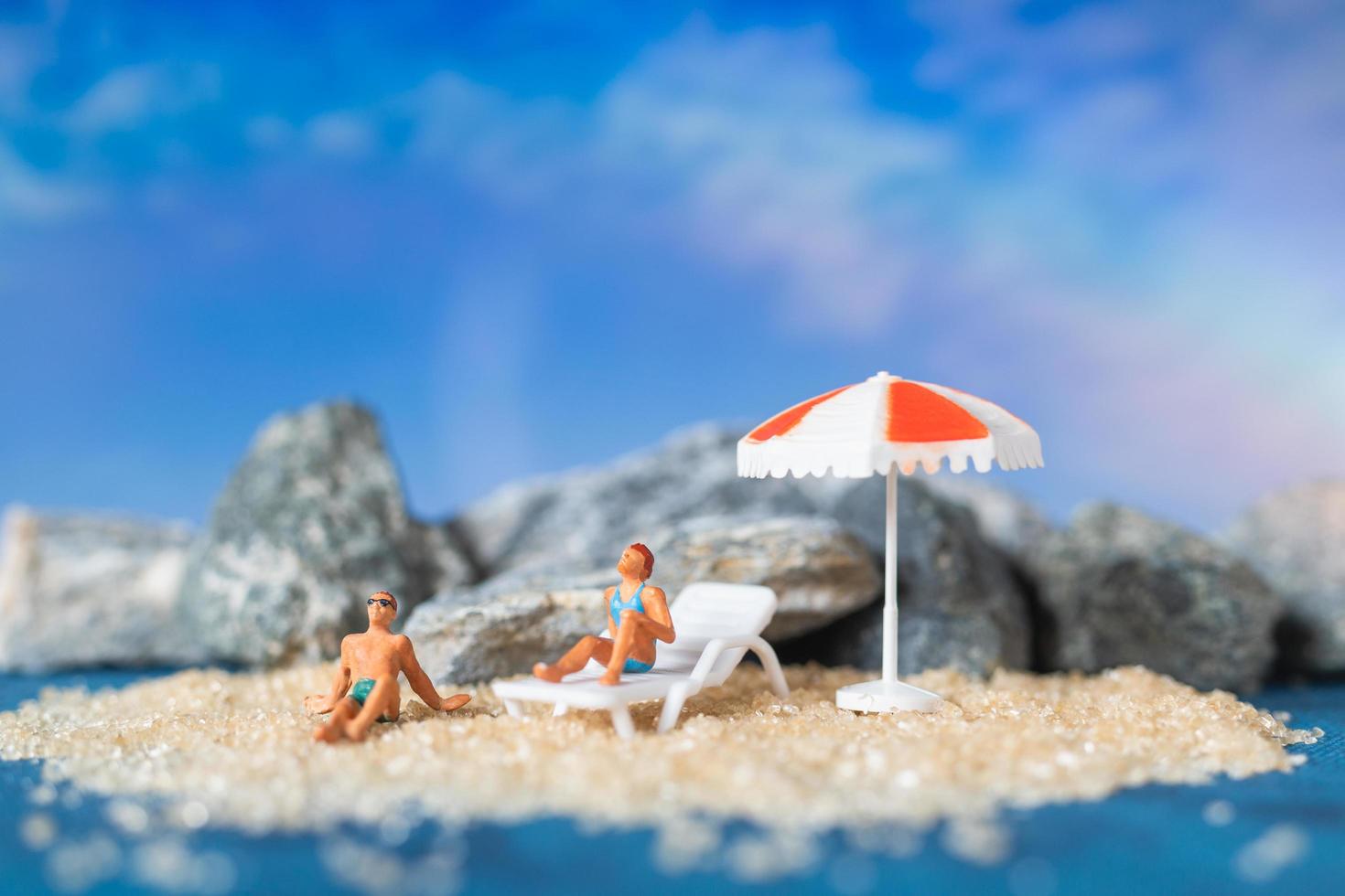 persone in miniatura che indossano costumi da bagno rilassante sulla spiaggia con uno sfondo blu, concetto di estate foto