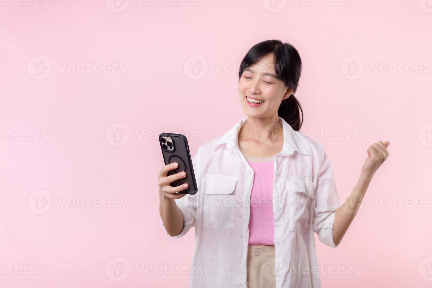 bella giovane asiatico donna mostrando successo, vittoria mano gesto mentre ricevente grande notizia a partire dal smartphone su rosa sfondo. contento tecnologia, mobile Telefono annuncio, in linea applicazione concetto foto