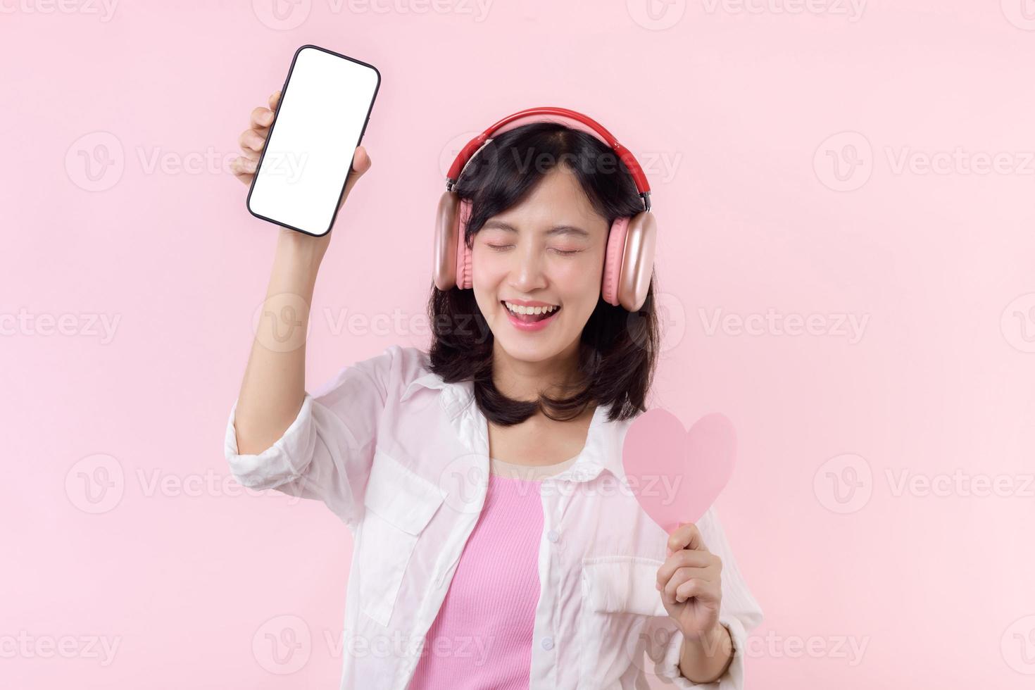 contento allegro sorridente asiatico donna con mano Tenere cuore e auricolari mostrando vuoto schermo mobile Telefono o nuovo smartphone musica applicazione annuncio pubblicitario modello isolato su rosa studio sfondo. foto