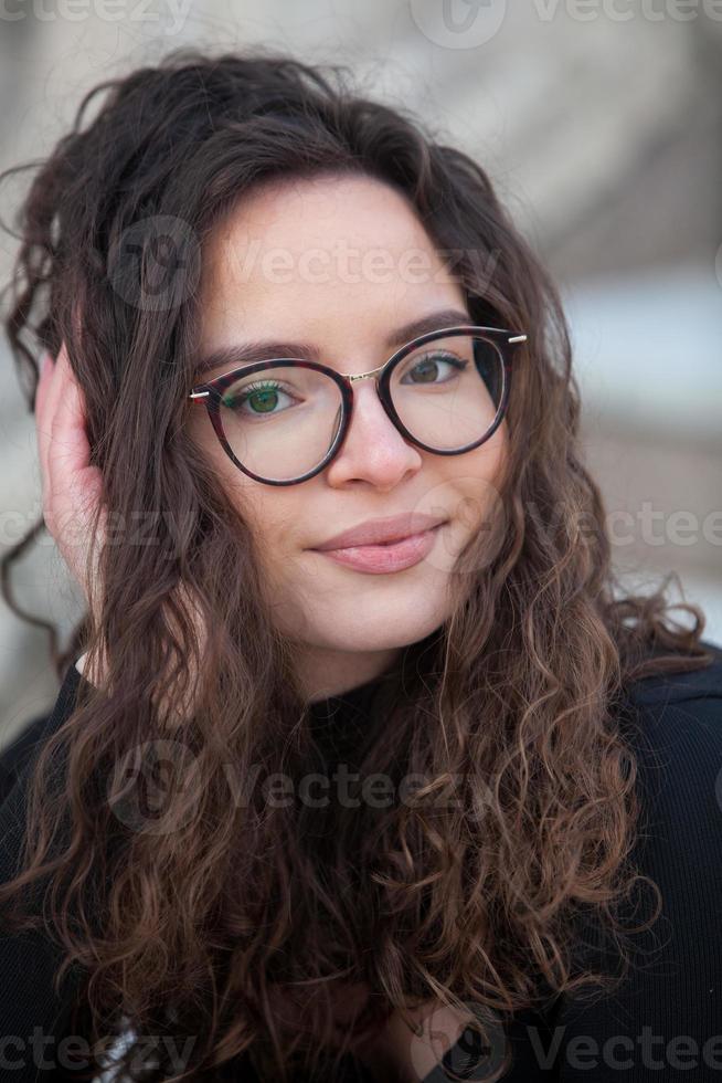 bellissimo giovane donna con brunetta Riccio capelli, ritratto nel occhio bicchieri godendo il sole nel il città. foto