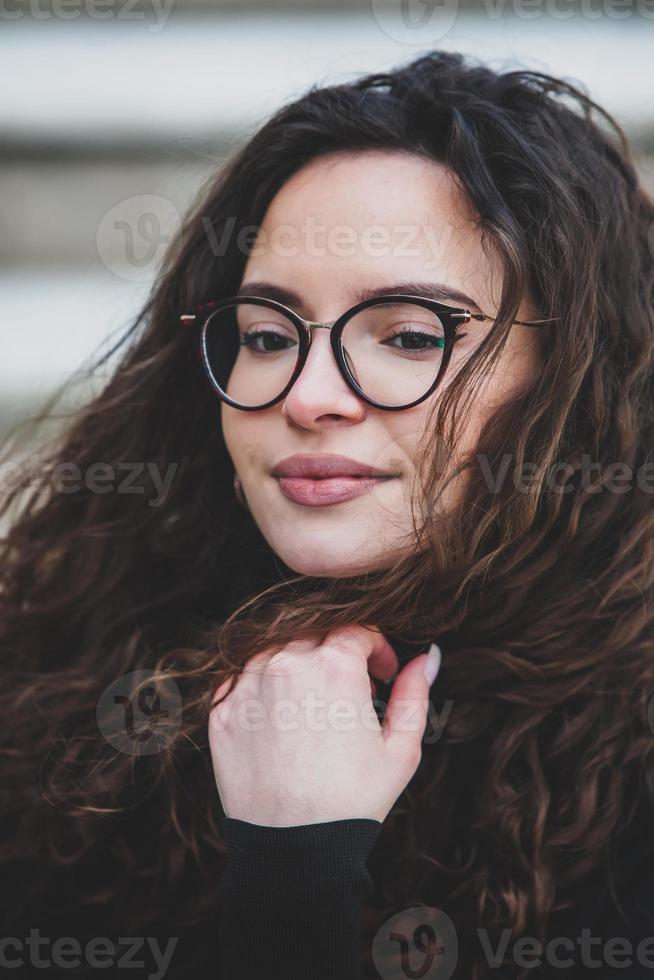 bellissimo giovane donna con brunetta Riccio capelli, ritratto nel occhio bicchieri godendo il sole nel il città. foto