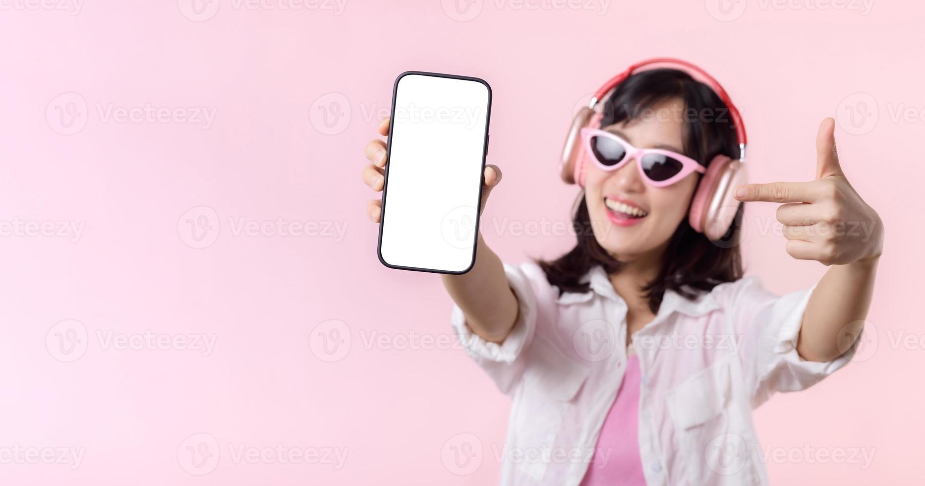 contento allegro sorridente asiatico donna con senza fili auricolari mostrando vuoto schermo mobile Telefono o nuovo smartphone musica applicazione annuncio pubblicitario modello isolato su rosa studio sfondo. foto