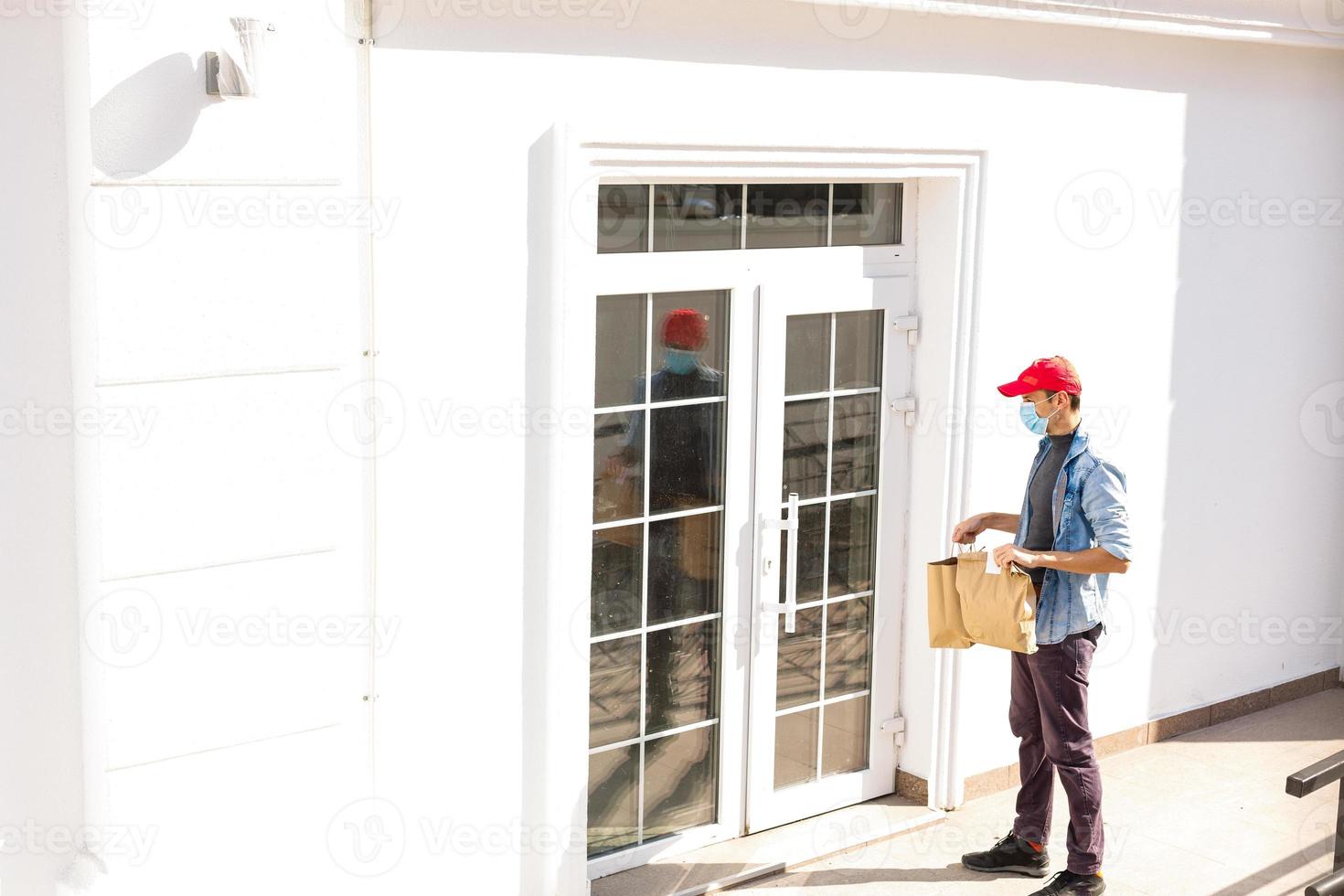 consegna uomo Tenere carta Borsa con cibo su bianca Ingresso di Casa sfondo , cibo consegna uomo nel protettivo maschera foto