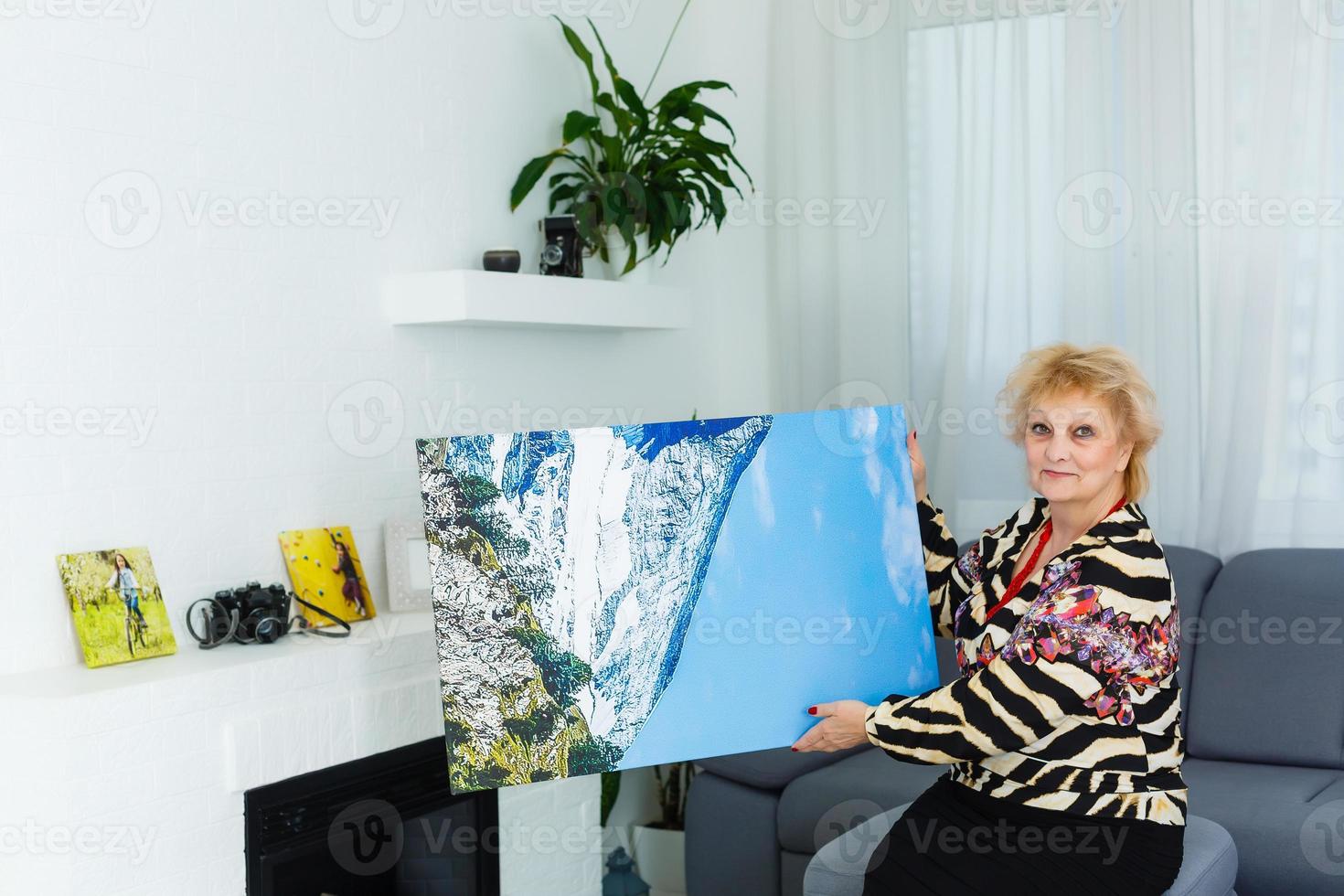 un' contento anziano bionda donna è Tenere un' grande parete foto tela a casa