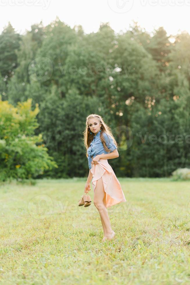 un' bellissimo giovane donna detiene estate scarpe nel sua mani e passeggiate a piedi nudi su il erba su un' estate giorno foto