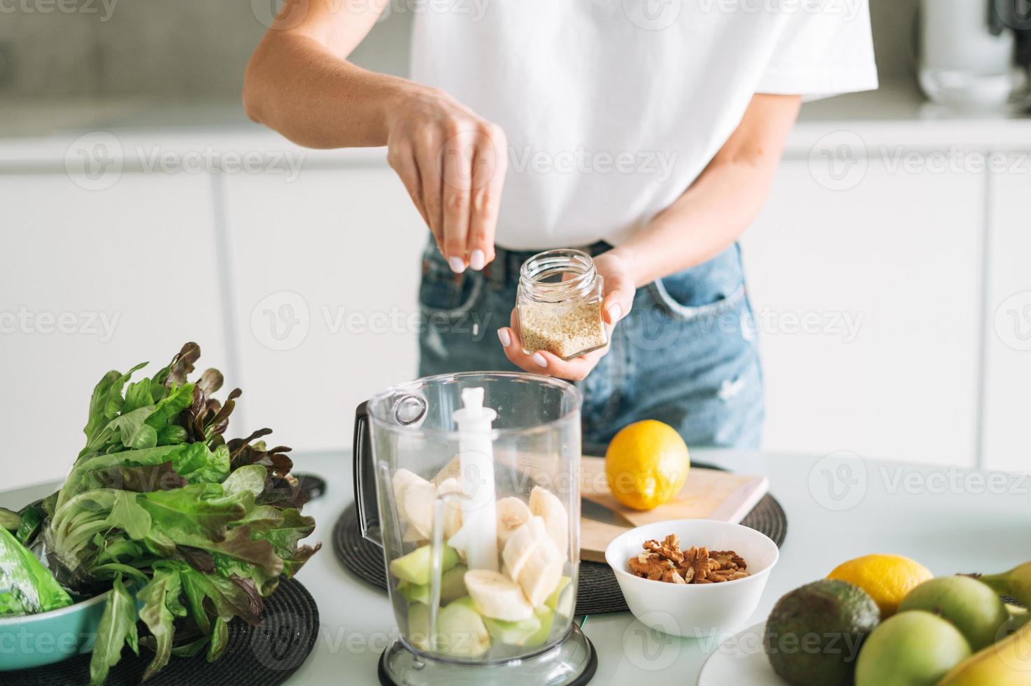 giovane sottile donna nel bianca maglietta e blu jeans cucinando frutta frullato con semi salutare cibo nel cucina a casa foto