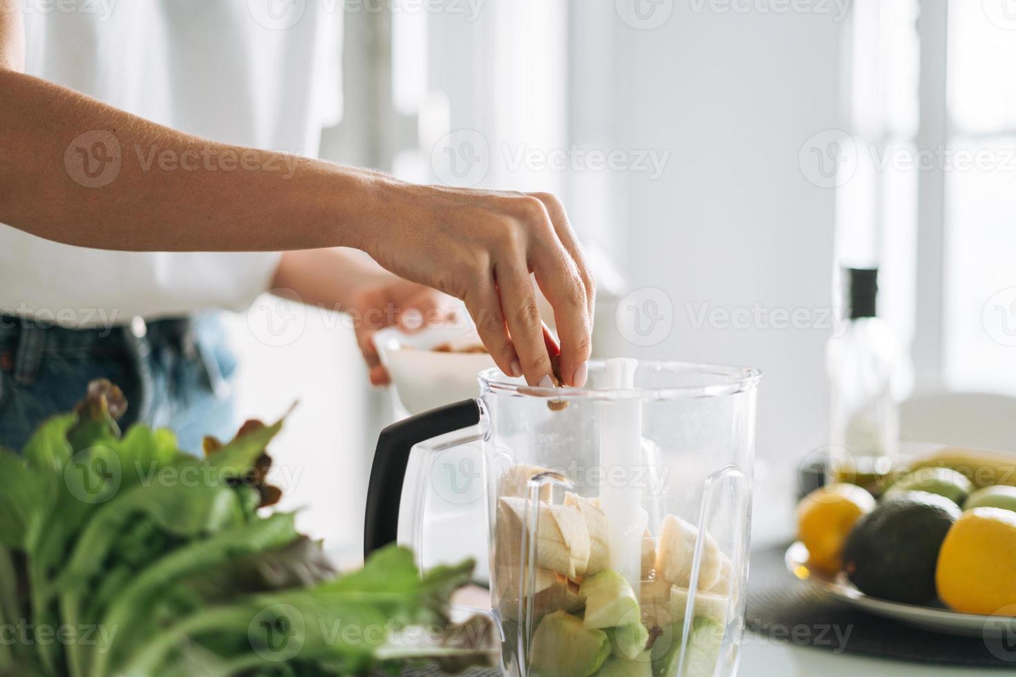 giovane sottile donna nel bianca maglietta e blu jeans cucinando frullato salutare cibo nel cucina a casa foto
