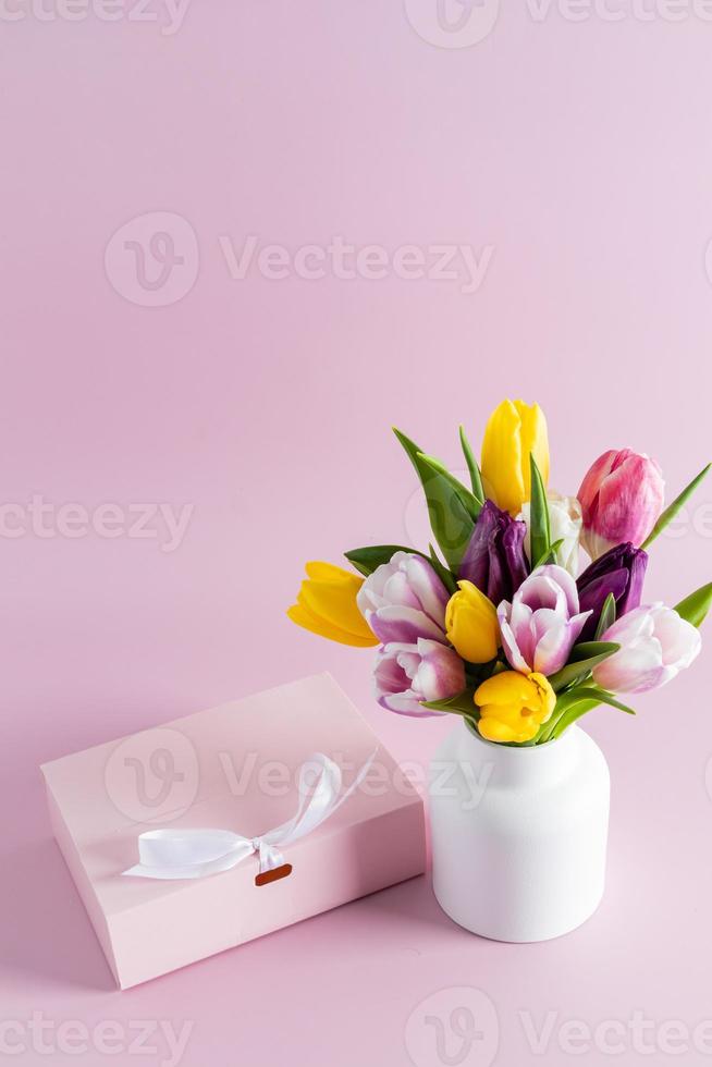 bellissimo mazzo di colorato tulipani nel un' bianca ceramica vaso su un' rosa sfondo con un' festivo regalo scatola. verticale Visualizza. un' copia spazio. foto