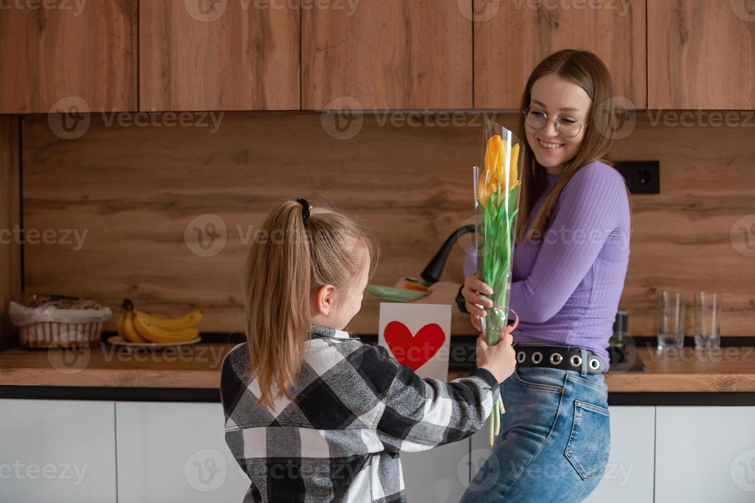 figlia si congratula mamma su La madre di giorno, carta con cuore e fiori. un' donna lavaggi piatti e è occupato con domestico lavoretti. foto