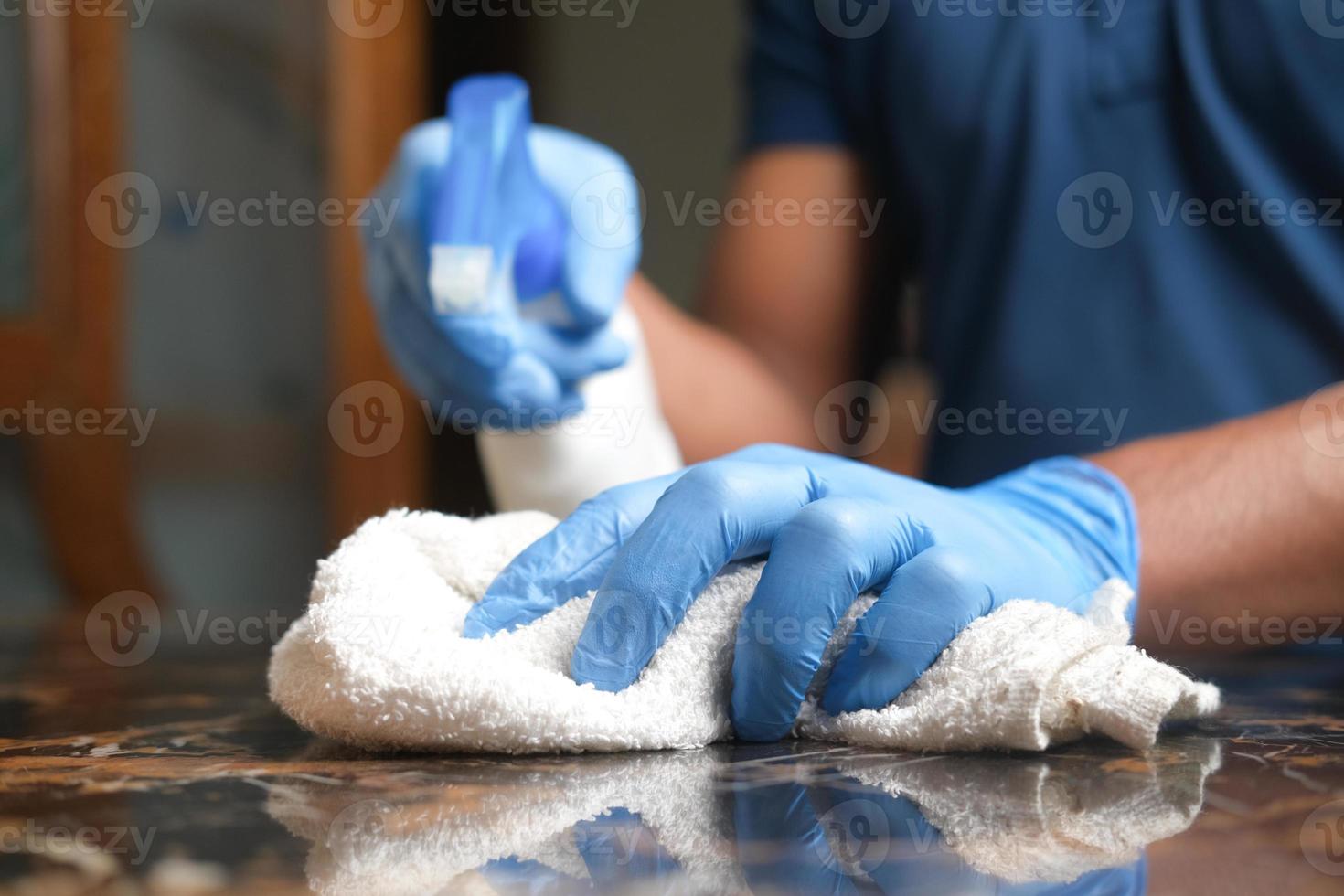 mano in guanti di gomma blu che tengono il flacone spray e puliscono un tavolo foto