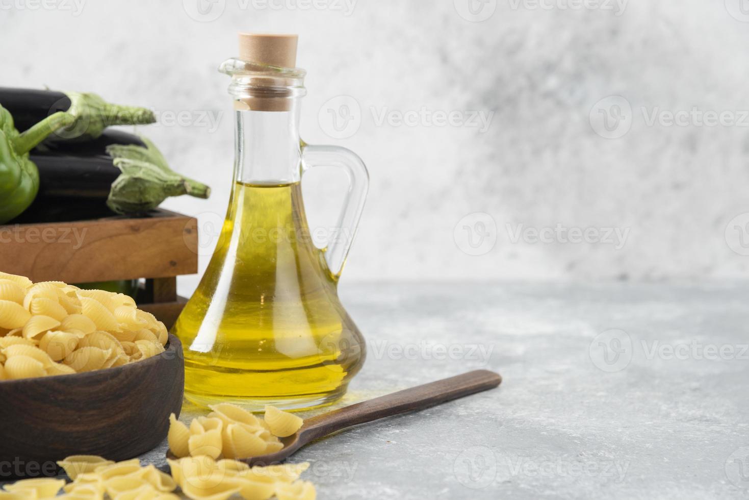 pasta cruda a forma di conchiglia con una bottiglia di vetro di olio e una scatola di legno piena di verdure fresche foto