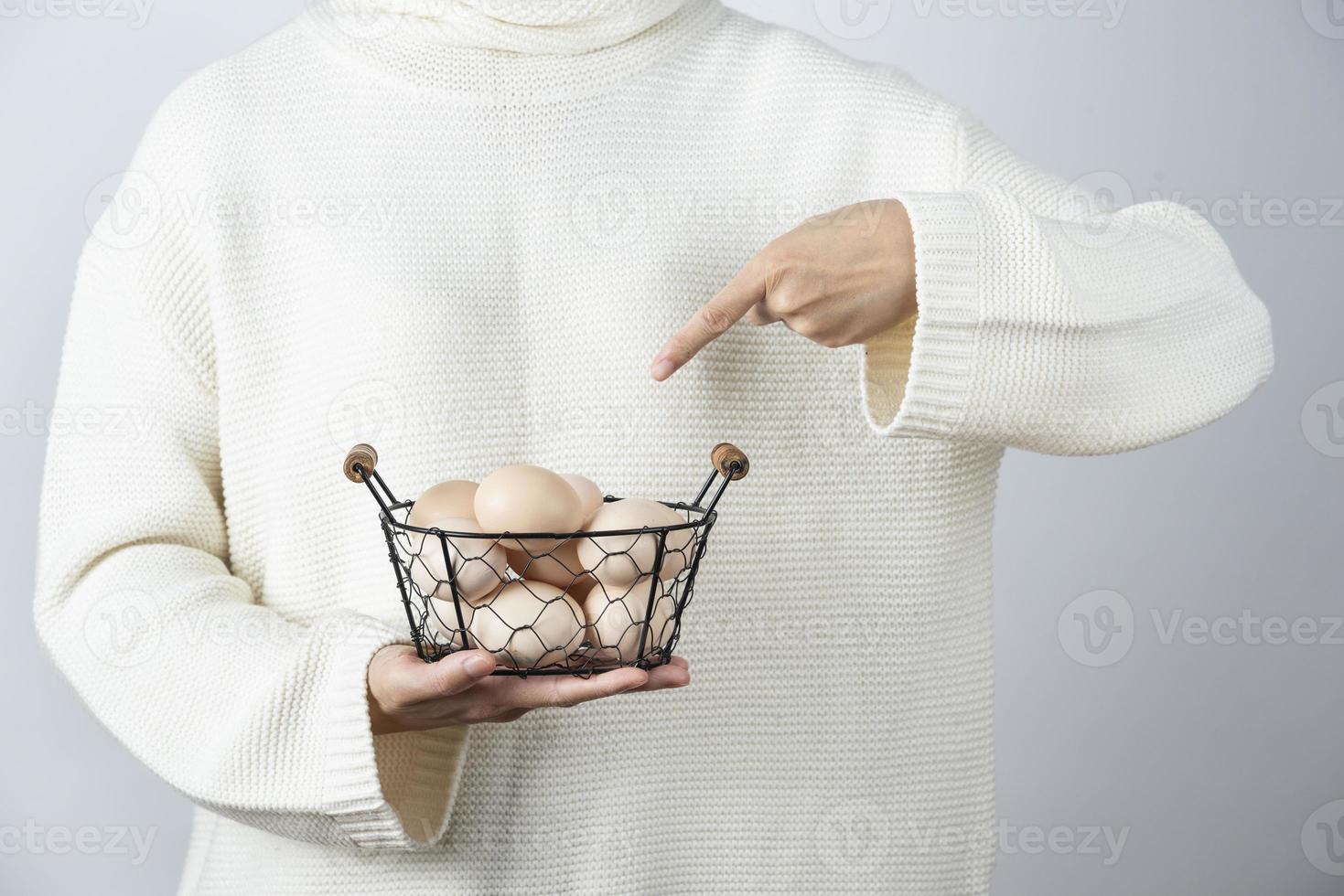 mani femminili che puntano a un cesto metallico pieno di uova di gallina crude contro un muro grigio foto