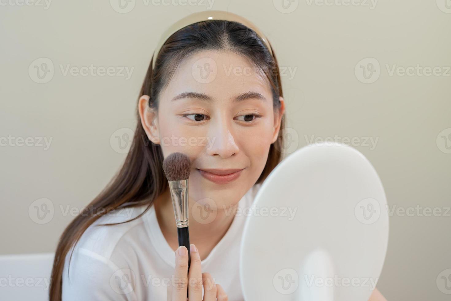contento routine bellezza concetto, bella asiatico giovane donna, ragazza rendere su viso di l'applicazione polvere fondazione di spazzola in giro viso, guardare a il specchio a casa. femmina Guarda con naturale moda stile. foto