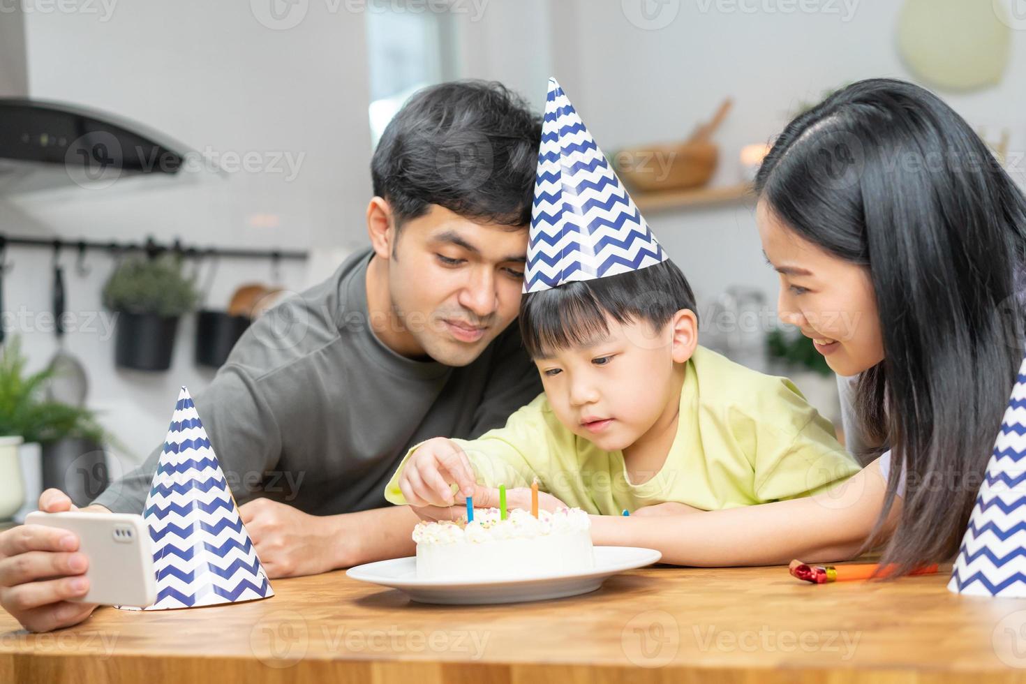 contento compleanno festa nel famiglia, asiatico giovane genitore padre, madre e poco carino ragazzo o bambino festeggiare, utilizzando smartphone autoscatto, prendere foto e video chiamata durante celebrazione insieme a casa.