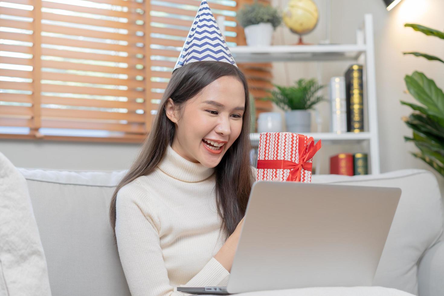 celebrare su giorno di contento compleanno stagione, bella asiatico giovane donna utilizzando il computer portatile computer, saluto su video chiamata durante sociale distanza, Tenere regalo scatola, contento per ottenere presente su divano, divano a casa. foto