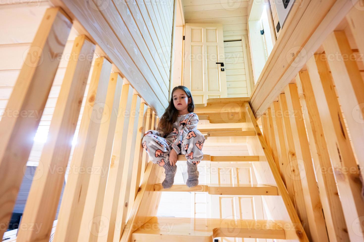 poco carino ragazza nel pigiama sedersi su le scale nel accogliente di legno minuscolo cabina Casa. vita nel campagna. foto