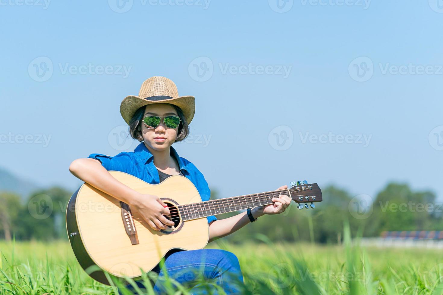 donne corto capelli indossare cappello e occhiali da sole sedersi giocando chitarra nel erba campo foto