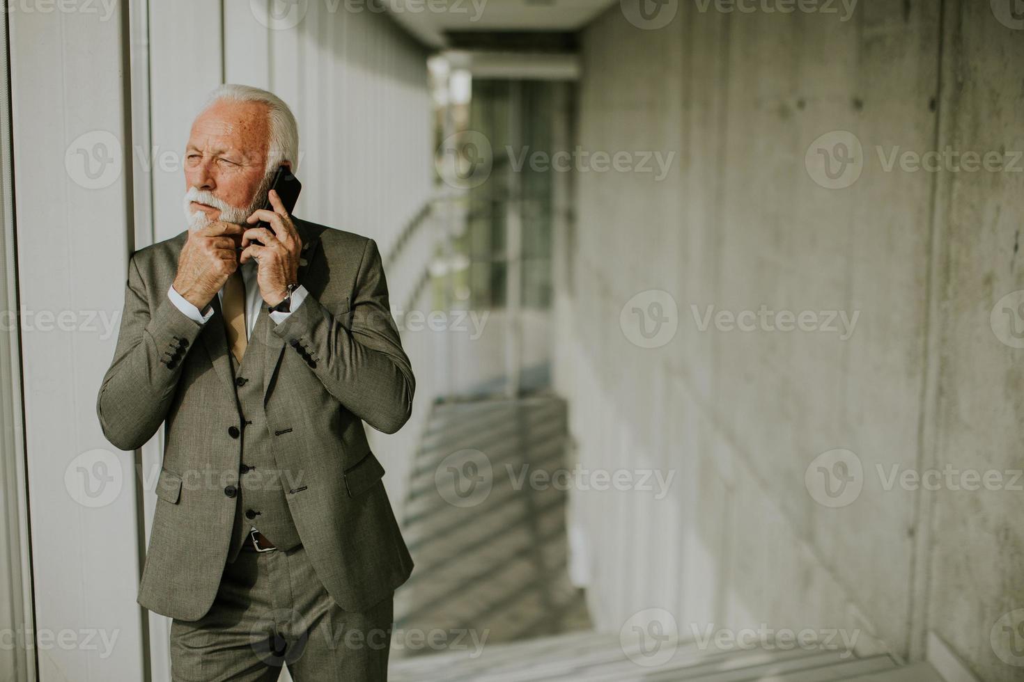 un' anziano attività commerciale uomo sta nel un ufficio corridoio, focalizzata su il suo mobile Telefono foto