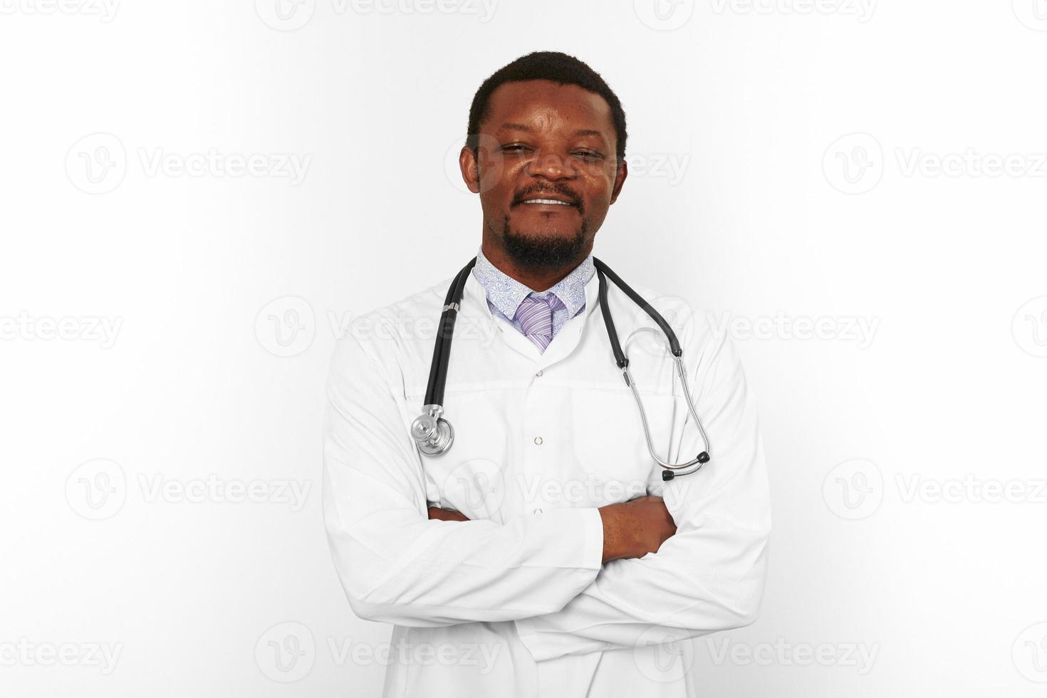 sorridente nero barbuto medico uomo attraversato braccia nel bianca accappatoio con stetoscopio, bianca sfondo foto