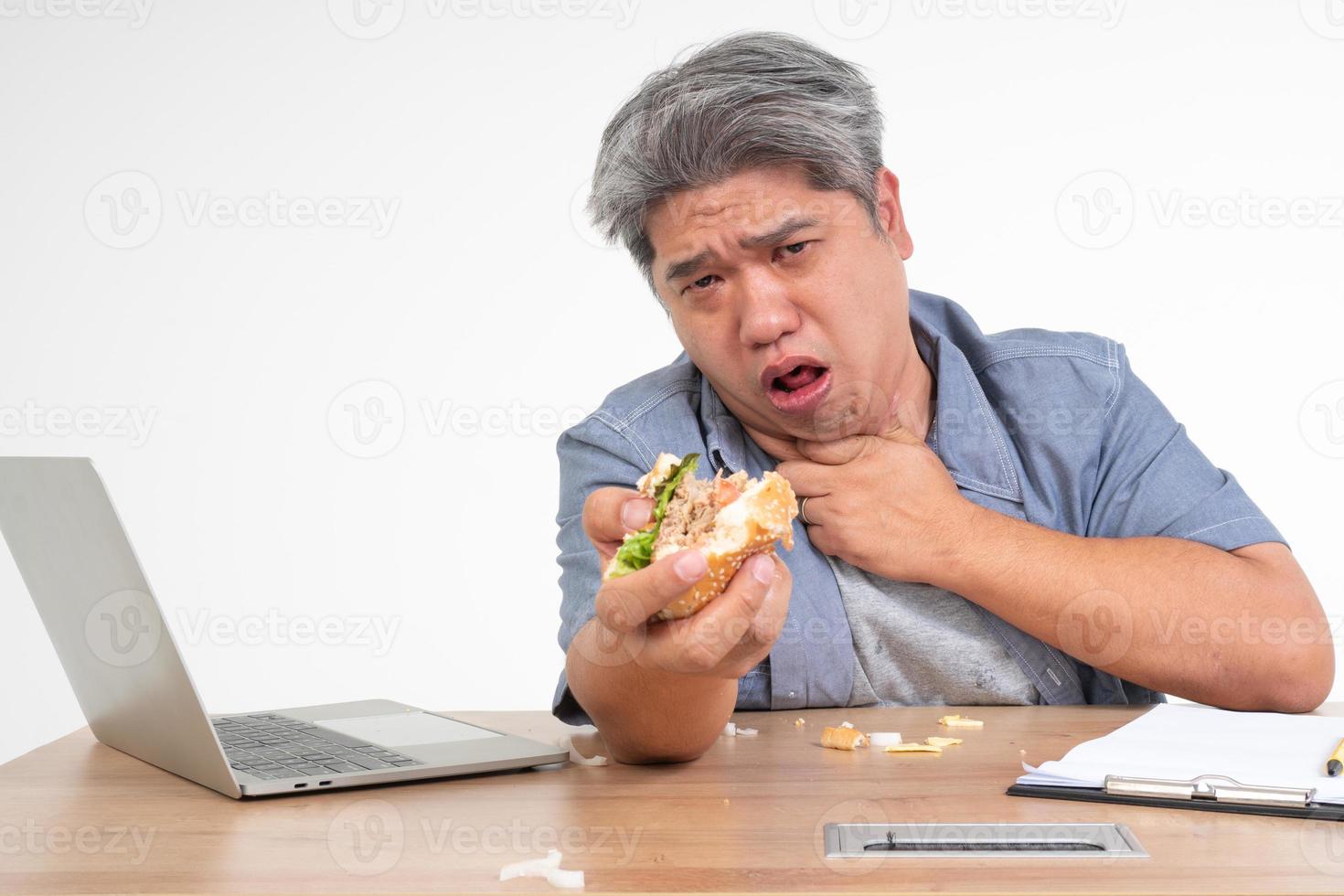 asiatico uomo Lavorando e mangiare un' hamburger su ufficio scrivania e Tenere il suo collo dopo soffocamento Alimenti. concetto di un' occupato uomo d'affari non può sinistra del lavoro equilibrio e non assunzione cura di Salute mangiare solo Rifiuto cibo foto
