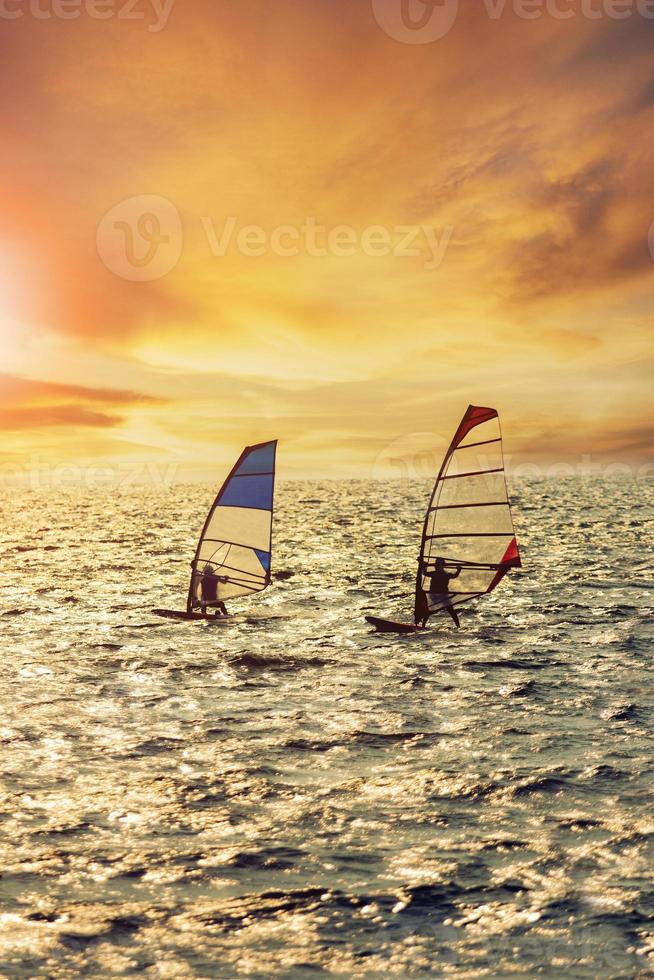 Due uomo giocando vento Surf al di sopra di affrettandosi mare contro bellissimo tramonto illuminazione foto
