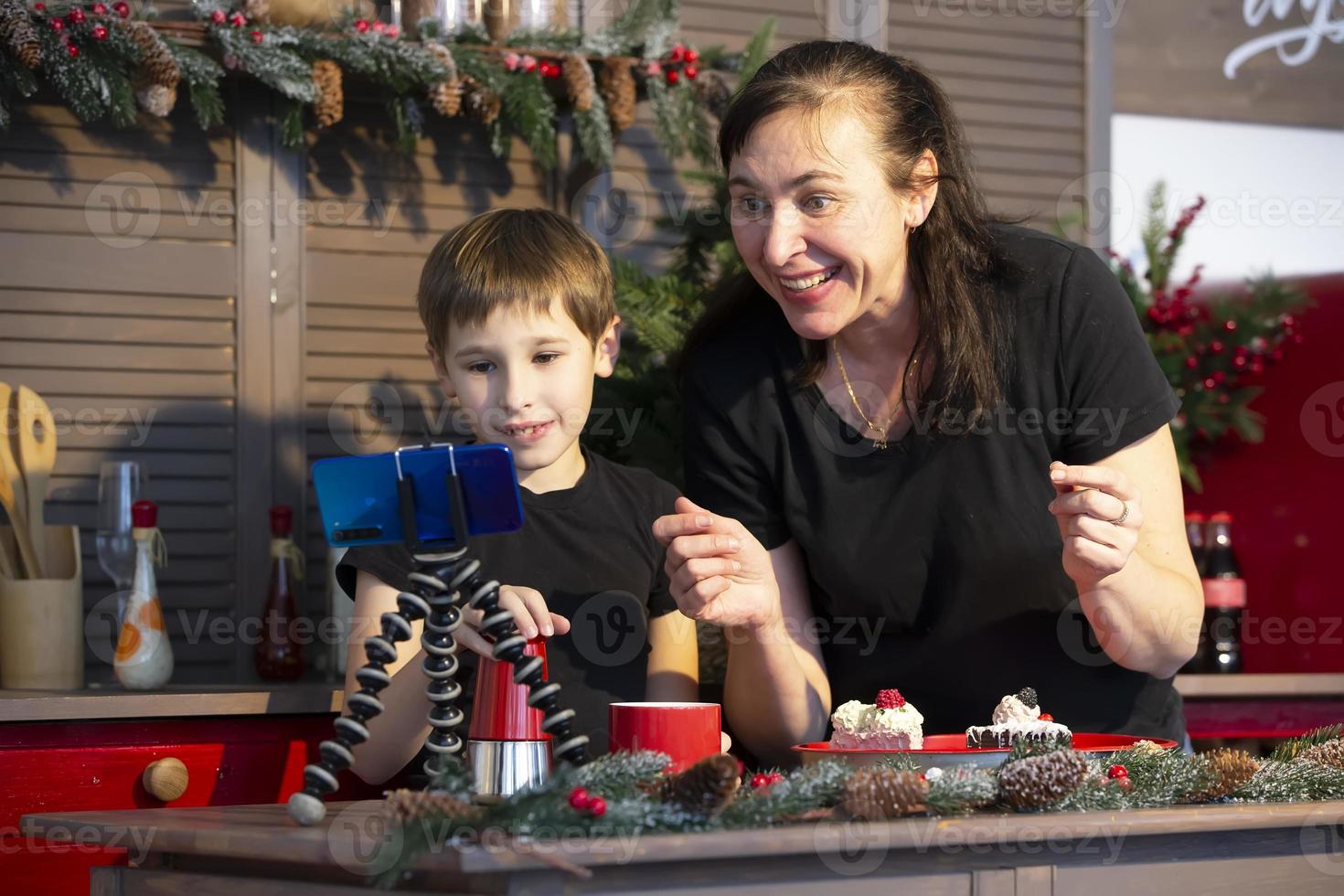 un' poco ragazzo e mamma siamo registrazione un' ruscello o video su Youtube su un' smartphone nel il casa cucina. foto