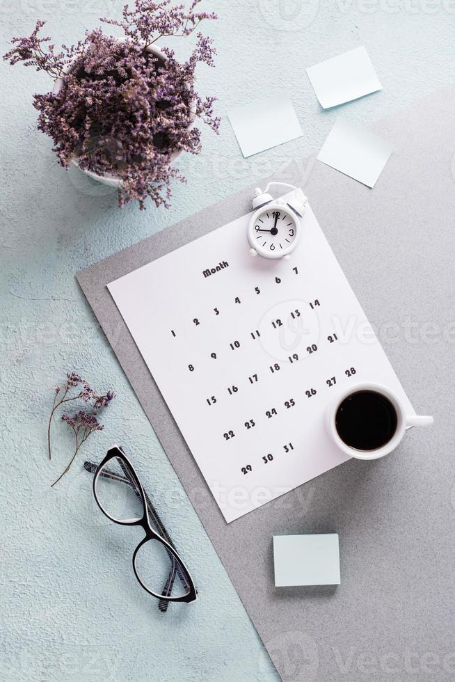 vuoto foglio di mensile calendario, occhiali, caffè tazza e allarme orologio su il desktop. tempo pianificazione e organizzazione. verticale Visualizza foto