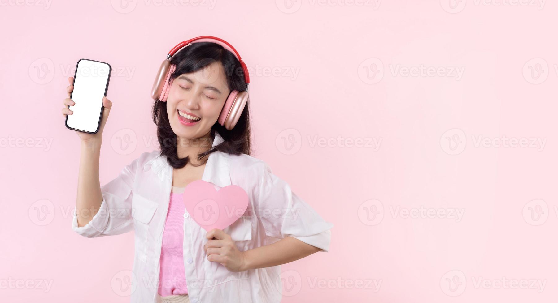 contento allegro sorridente asiatico donna con mano Tenere cuore e auricolari mostrando vuoto schermo mobile Telefono o nuovo smartphone musica applicazione annuncio pubblicitario modello isolato su rosa studio sfondo. foto