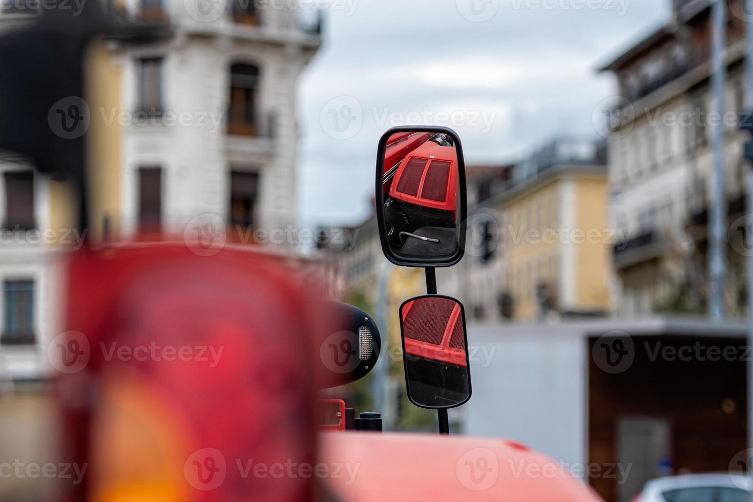 primo piano dello specchietto laterale del trattore su sfondo sfocato della città. lo specchio mostra il cofano rosso del trattore. foto