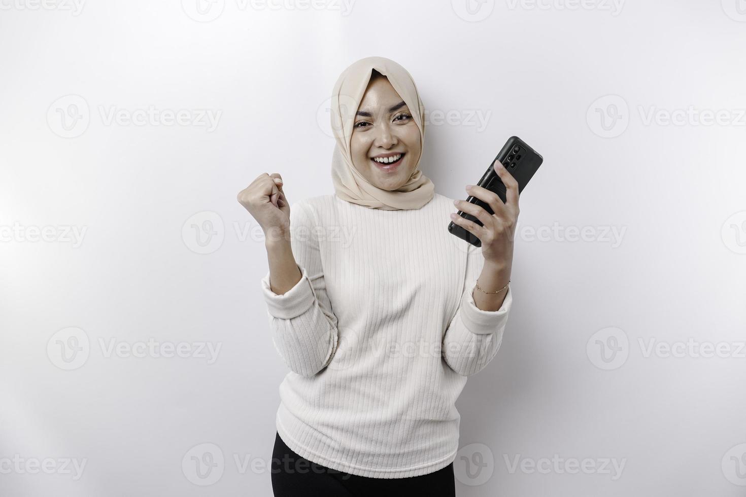 un' giovane asiatico musulmano donna con un' contento riuscito espressione indossare un' hijab e Tenere smartphone isolato di bianca sfondo foto
