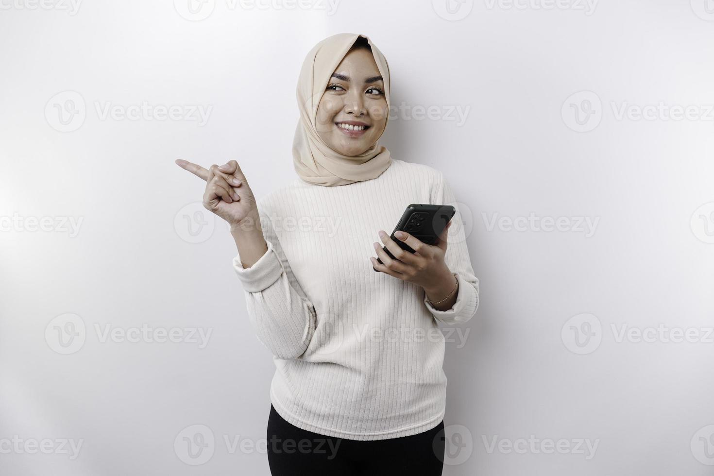 eccitato asiatico donna indossare hijab puntamento a il copia spazio accanto sua mentre Tenere sua Telefono, isolato di bianca sfondo foto