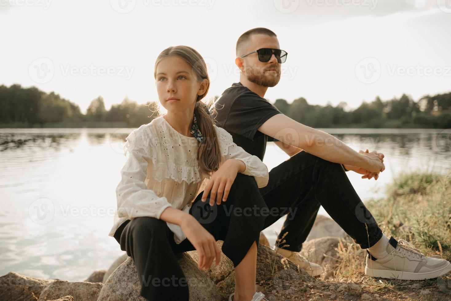 una giovane ragazza posa sulle pietre con il padre con la barba e gli occhiali da sole sulla costa del lago foto