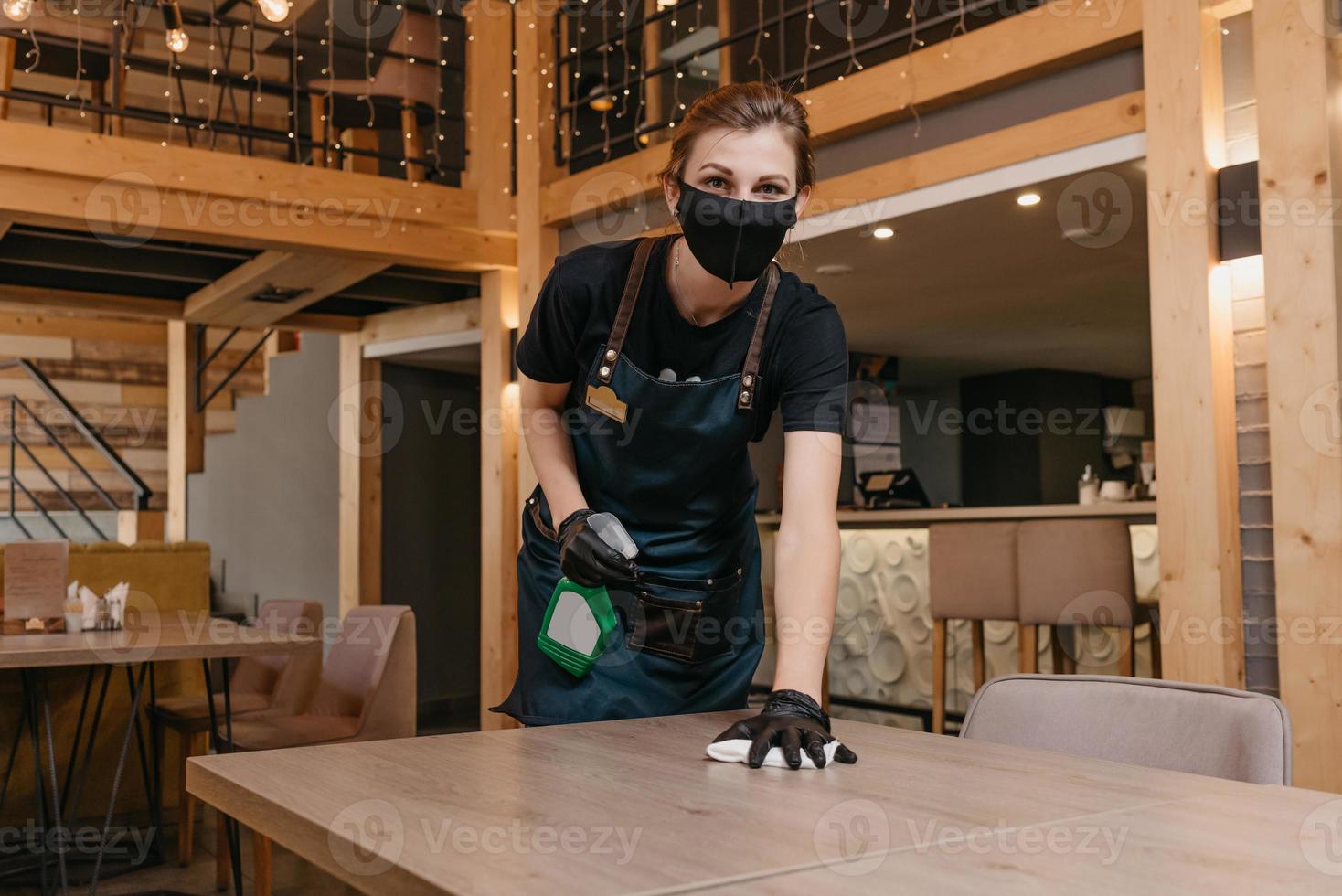 una cameriera gentile che indossa una maschera medica nera e guanti medici usa e getta tiene in mano una bottiglia con disinfettante e pulisce i tavoli con uno straccio in un ristorante foto