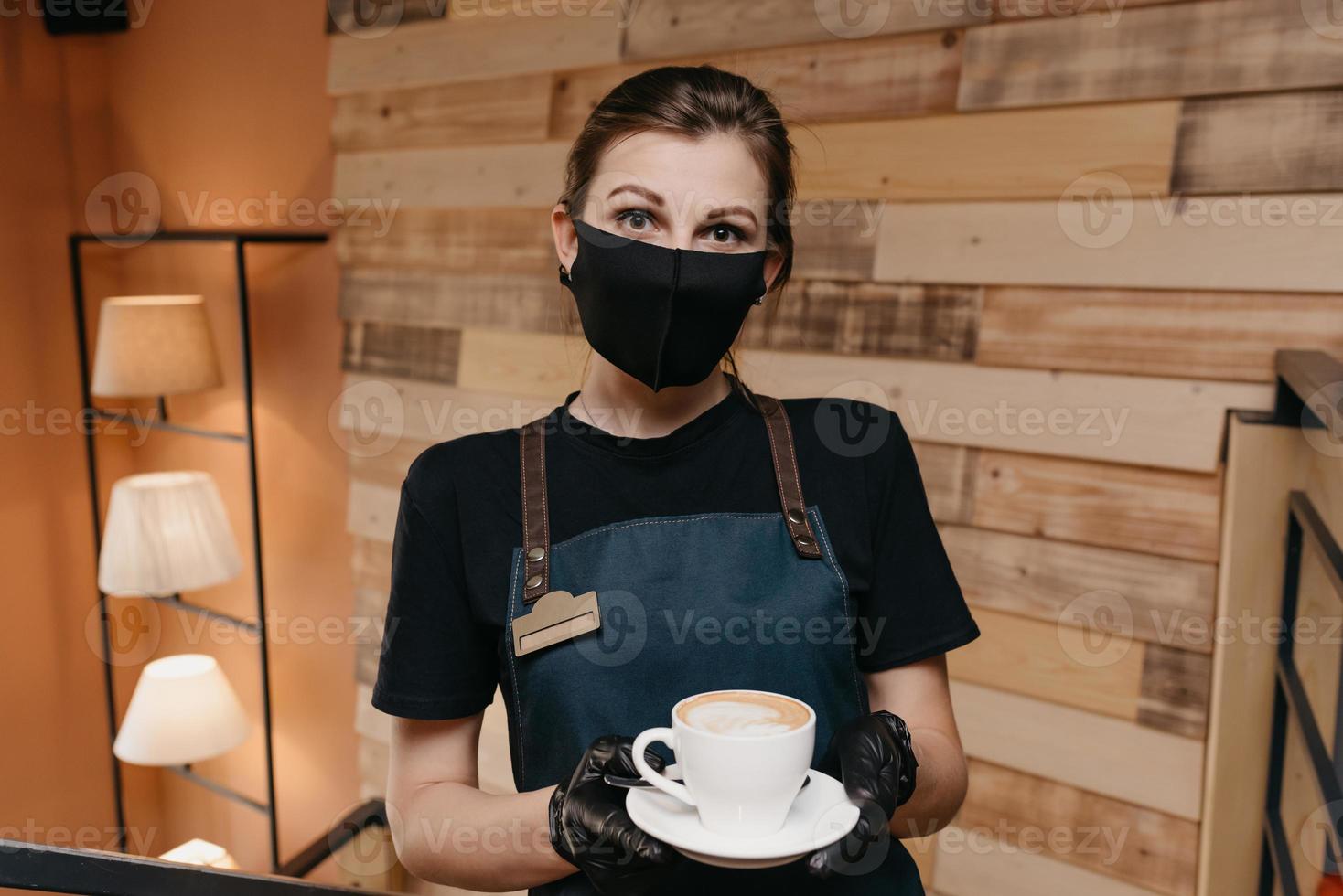 una cameriera che indossa una maschera nera e guanti usa e getta serve i clienti in un bar foto