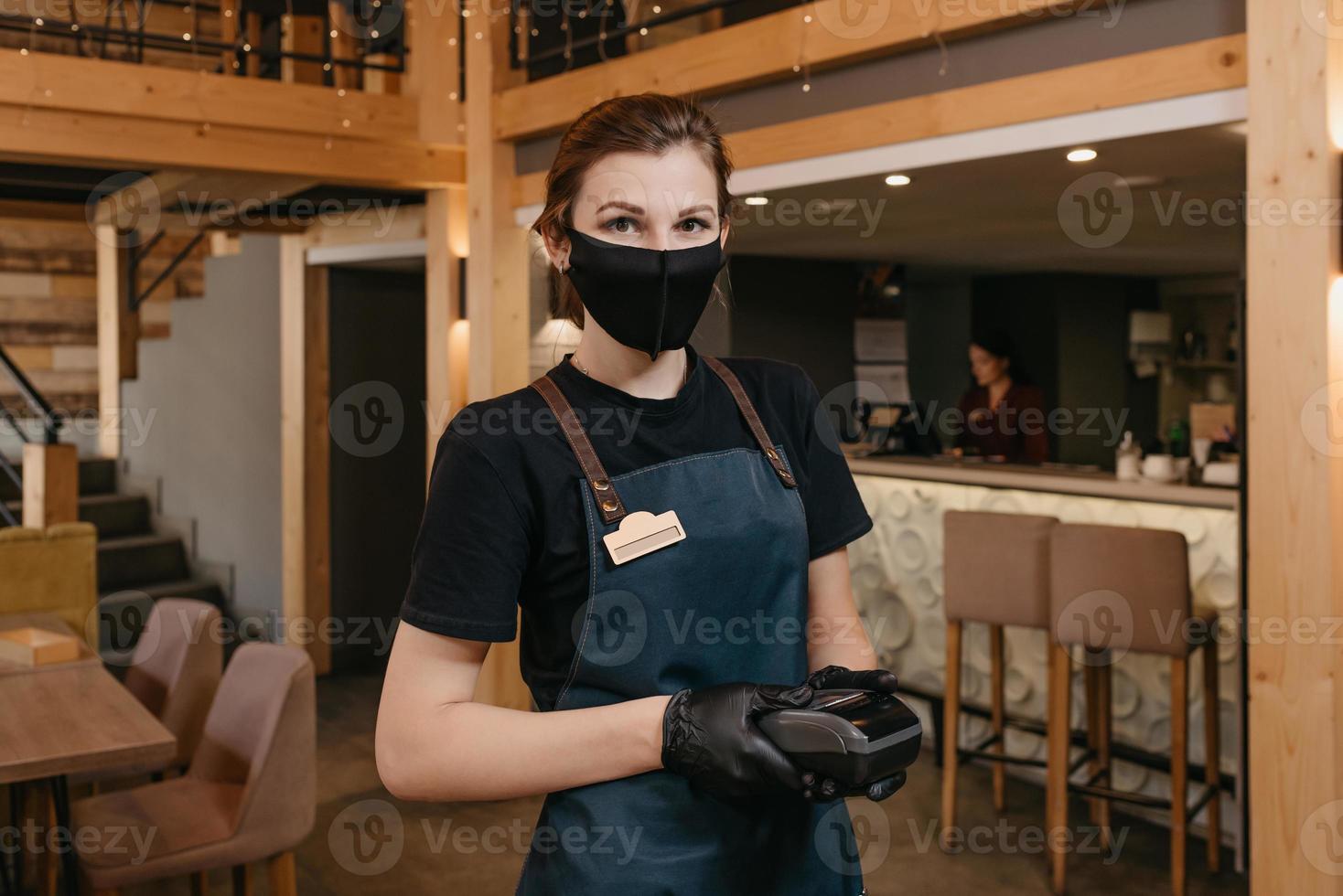 una direttrice di un ristorante indossa una maschera nera e guanti usa e getta in possesso di un terminale di pagamento in un ristorante foto