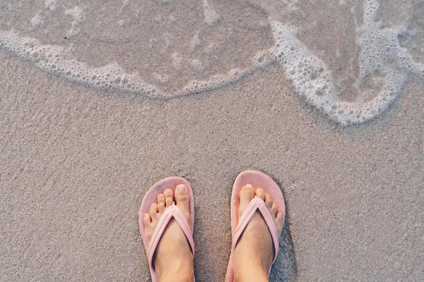 vista dall'alto dei piedi di una donna che indossa pantofole in piedi su una spiaggia di sabbia con le onde del mare foto
