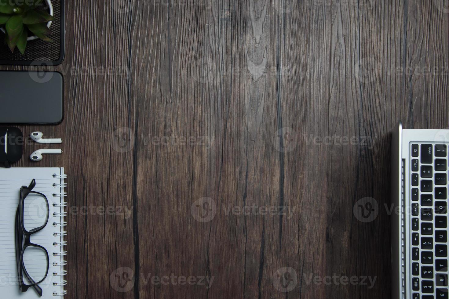 di legno ufficio scrivania con computer portatile, vuoto taccuino, cordless auricolari e occhiali, buio sfondo, di legno sfondo, superiore Visualizza con copia spazio, vuoto spazio. foto