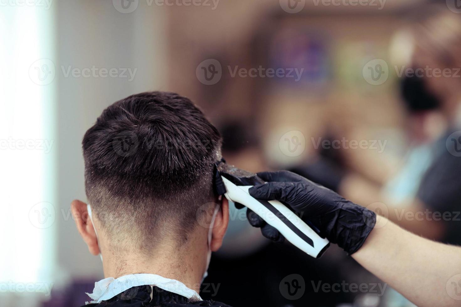 un' parrucchiere con sicurezza le misure per covid19, detiene un' rasoio e un' taglio di capelli per un' uomo, parla a un' distanza, tagli il suo capelli con gomma da cancellare guanti foto