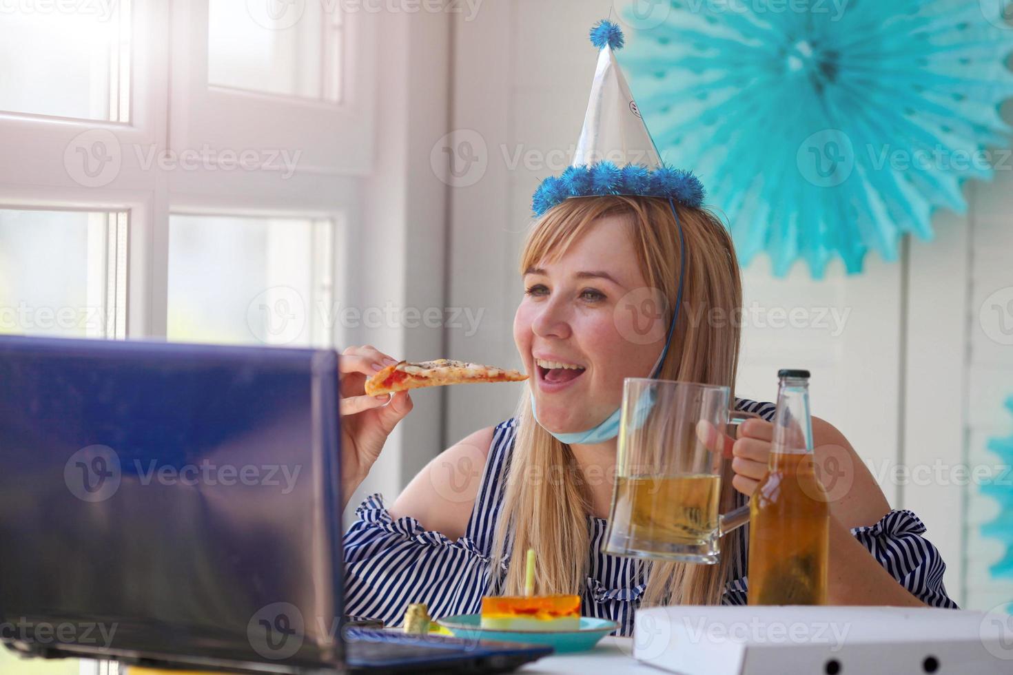 bellissimo donna nel un' medico maschera è seduta nel davanti di un' il computer portatile e mangiare Pizza. celebra il suo compleanno al di sopra di il Internet nel quarantena volta. foto