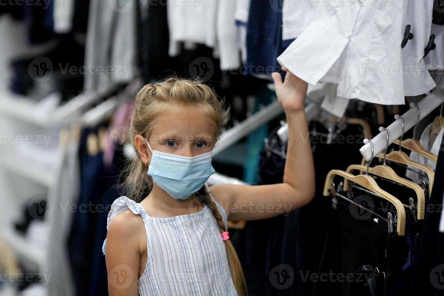 poco ragazza nel un' medico maschera sceglie scuola uniforme negozio. preparazione per scuola dopo quarantena covid - 19. protettivo maschera per il prevenzione di coronavirus foto