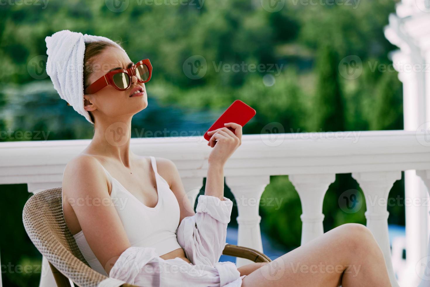 ritratto di giovane donna utilizzando mobile Telefono mentre seduta su balcone con un' bellissimo natura Visualizza pigro mattina foto