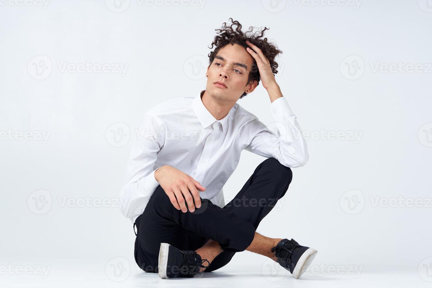 un' tipo nel i pantaloni nel scarpe da ginnastica e un' camicia si siede su il pavimento e gesti con il suo mani nel pieno crescita foto