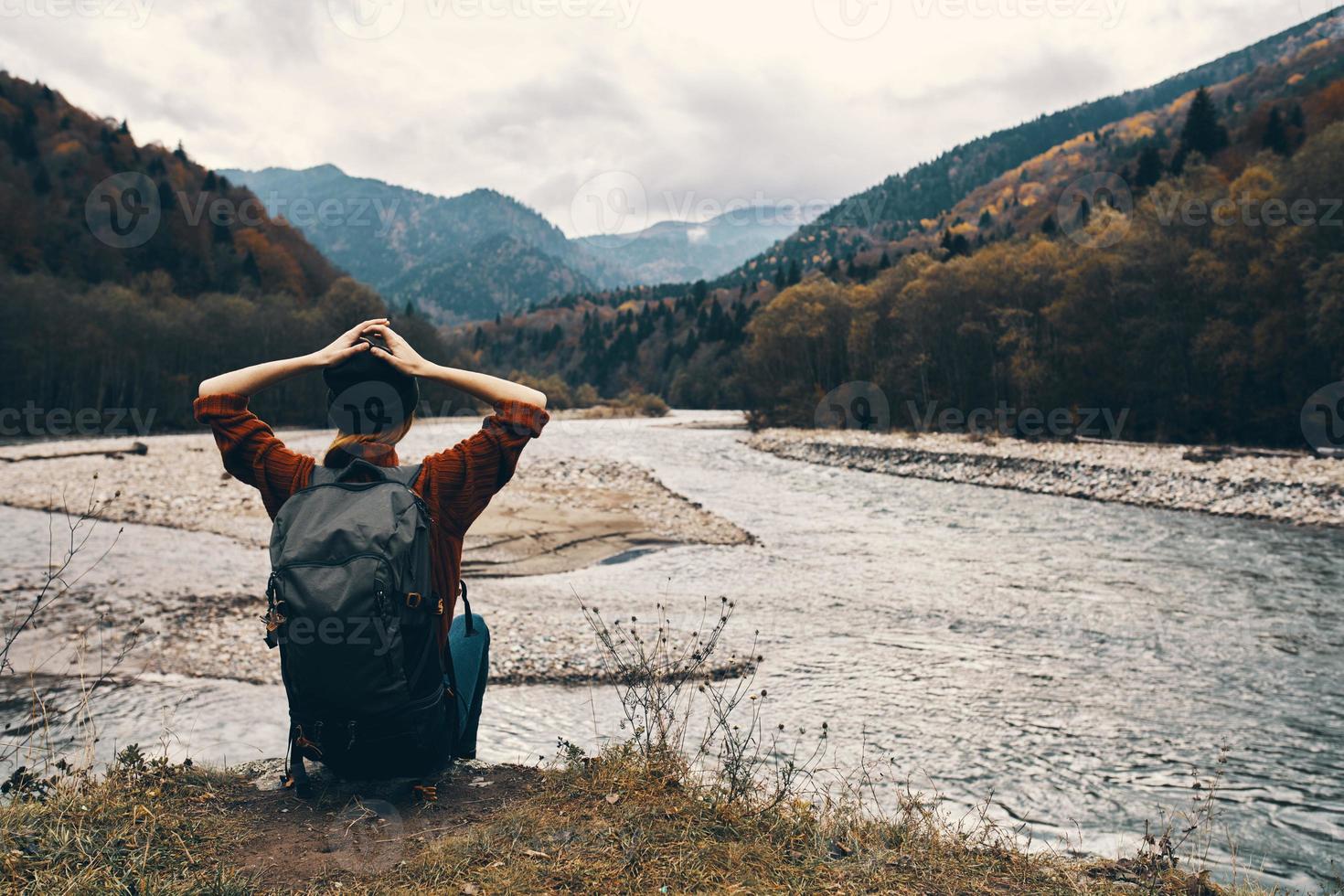 donna escursionista si siede su il fiume banca e ammira il montagna paesaggio nel il distanza indietro Visualizza foto