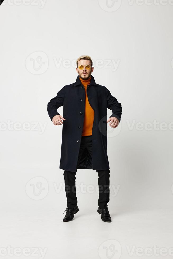 alla moda uomo nel un' cappotto detiene il suo mani su il suo cintura e nero i pantaloni modello leggero sfondo foto
