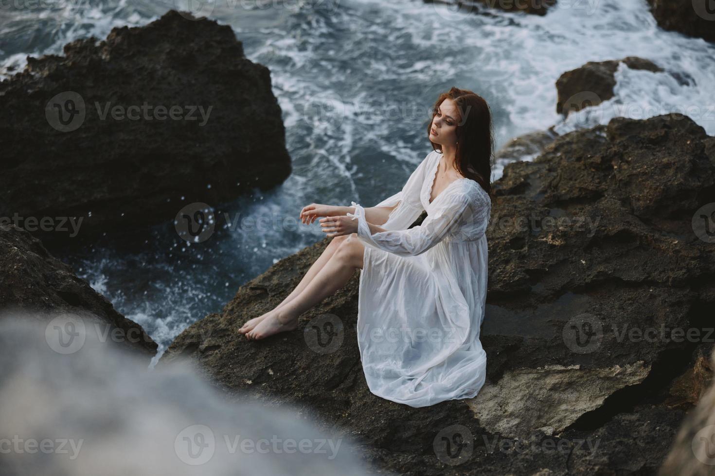 bella donna con bagnato capelli nel un' bianca vestito su il costa si siede su il rocce foto