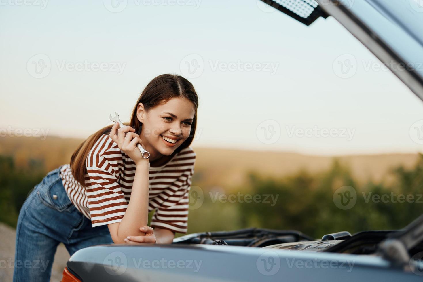 un' donna con un' chiave inglese con un' Sorridi felicemente sembra sotto il Aperto cappuccio di sua auto e riparazione esso a partire dal un' ciglio della strada abbattersi su un' strada viaggio solo foto