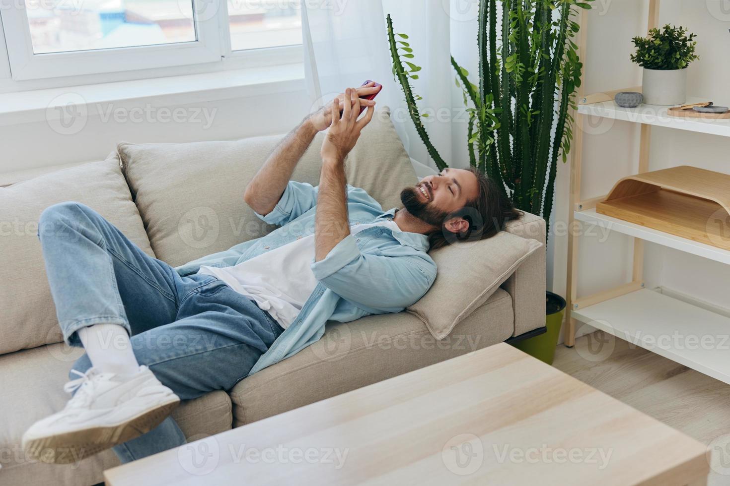 un' uomo con un' barba bugie su il divano durante il giorno a casa e sembra a il suo Telefono rilassante su il suo giorno spento, un' uomo gioco d'azzardo su il azione mercato in linea su il suo Telefono foto